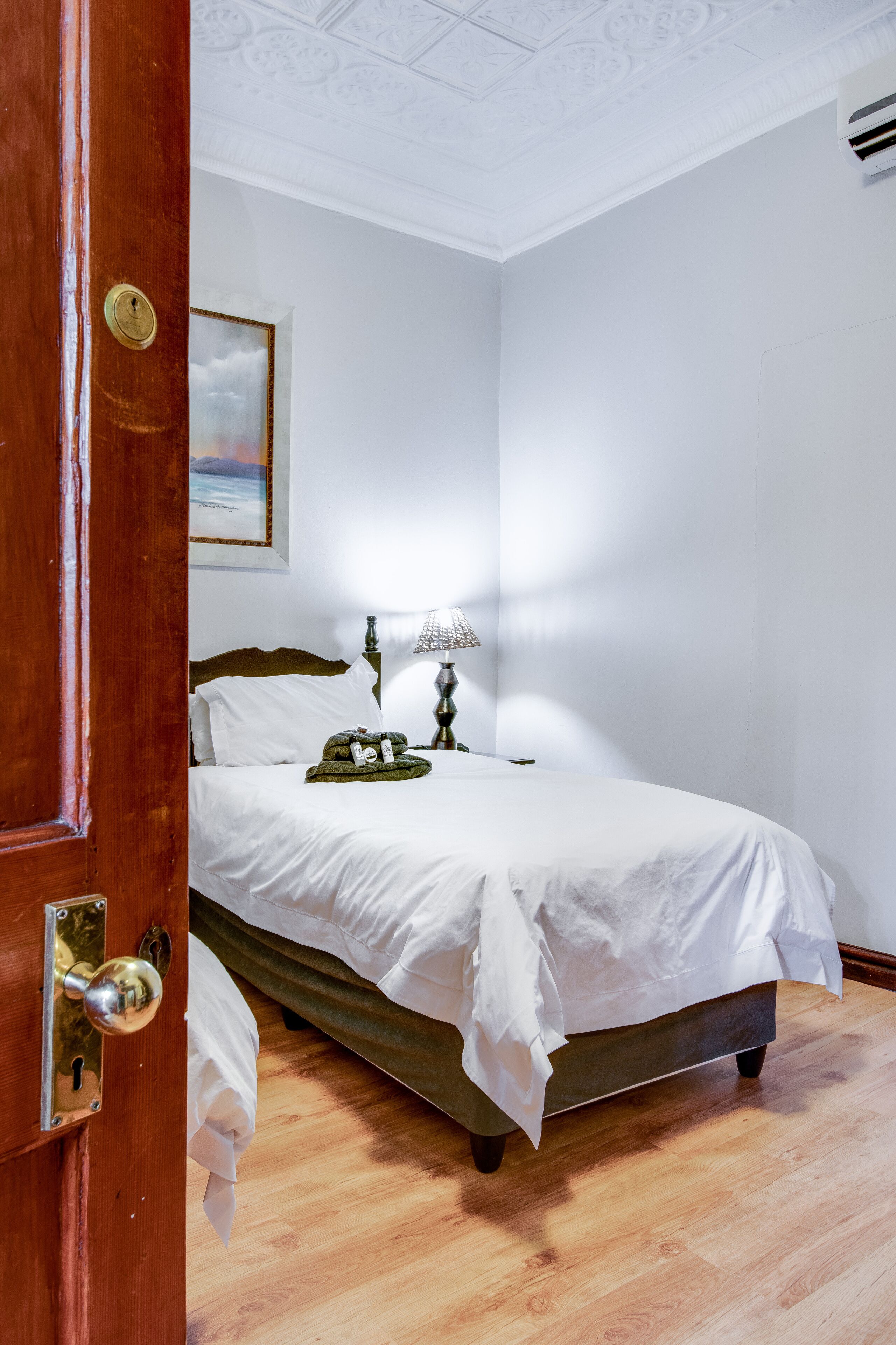 Une chambre avec une porte en bois, un lit recouvert de draps blancs, une lampe et un tableau accroché au mur.