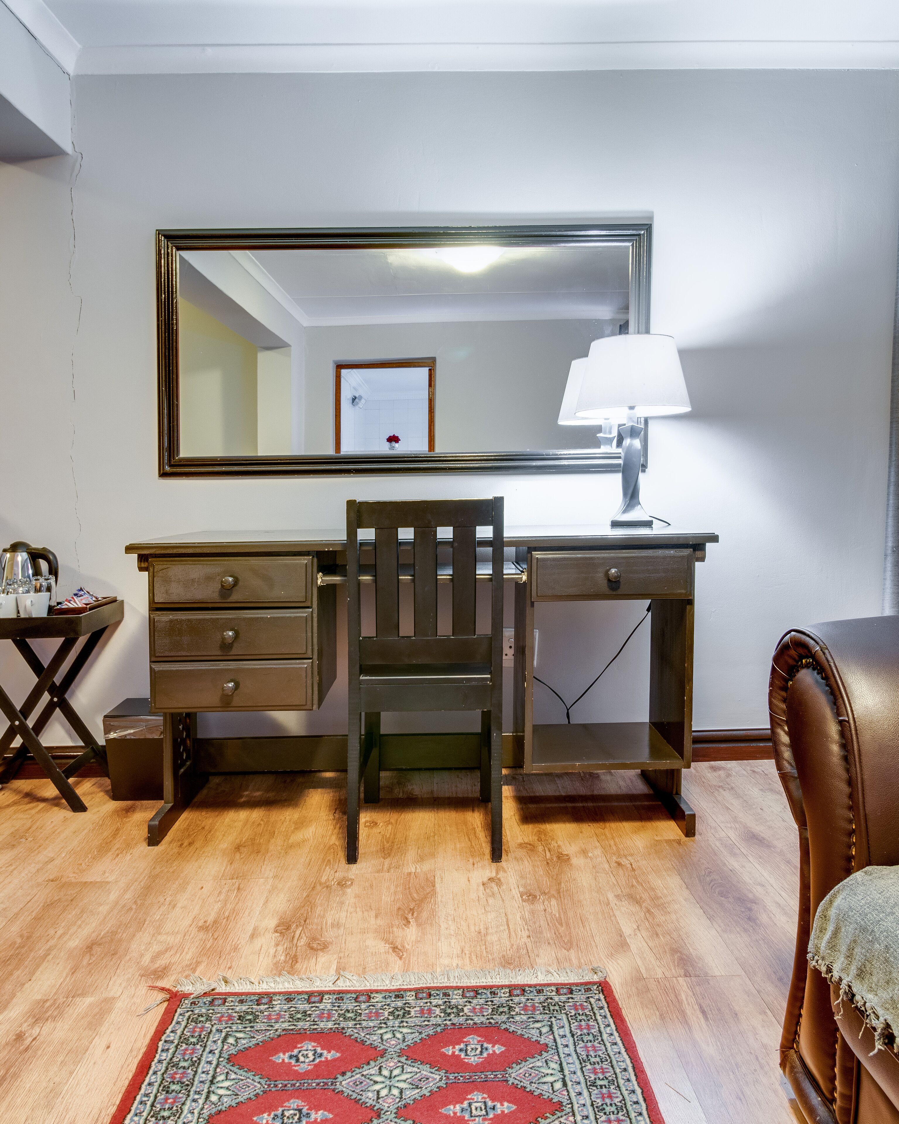 A room with a wooden desk, a chair, a mirror, a lamp, and a patterned rug.