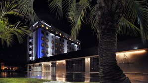 Un hôtel doté d’une façade moderne, d’une enseigne lumineuse et d’un espace extérieur bien entretenu, agrémenté de palmiers et d’un sentier.