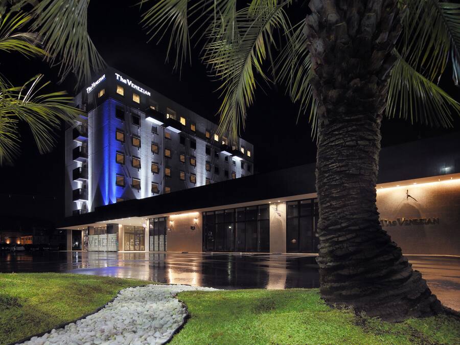Hotel amb façana moderna, senyalització il·luminada i zona exterior ben cuidada amb palmeres i un camí.