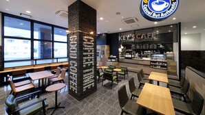 L’intérieur d’un café moderne, avec des espaces aménagés pour s’asseoir, un pilier en briques bien visible et une grande fenêtre qui laisse entrer la lumière naturelle.