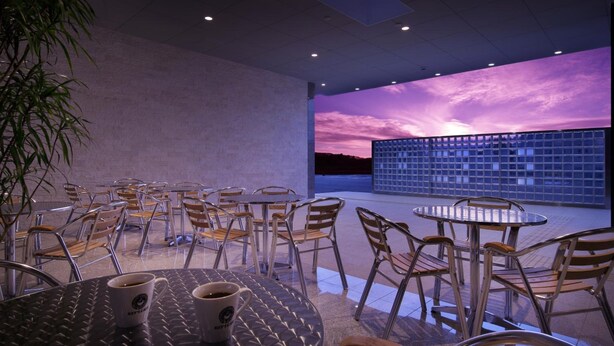Un espace de détente intérieur moderne, avec des tables et des chaises, une grande fenêtre donnant sur le ciel et une plante en pot.