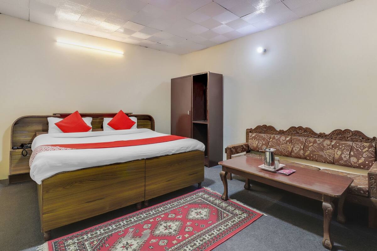 Un dormitorio con cama de madera, colcha roja y blanca, almohada decorativa roja, mesita de noche de madera, armario de madera, sofá estampado y una pequeña mesa con bandeja y tetera.