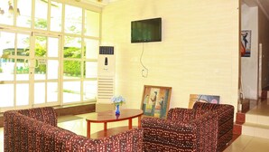 A room with a large window, a flat-screen TV mounted on the wall, a small table with a vase, and patterned sofas. - Carlcon Hotel (Calabar)