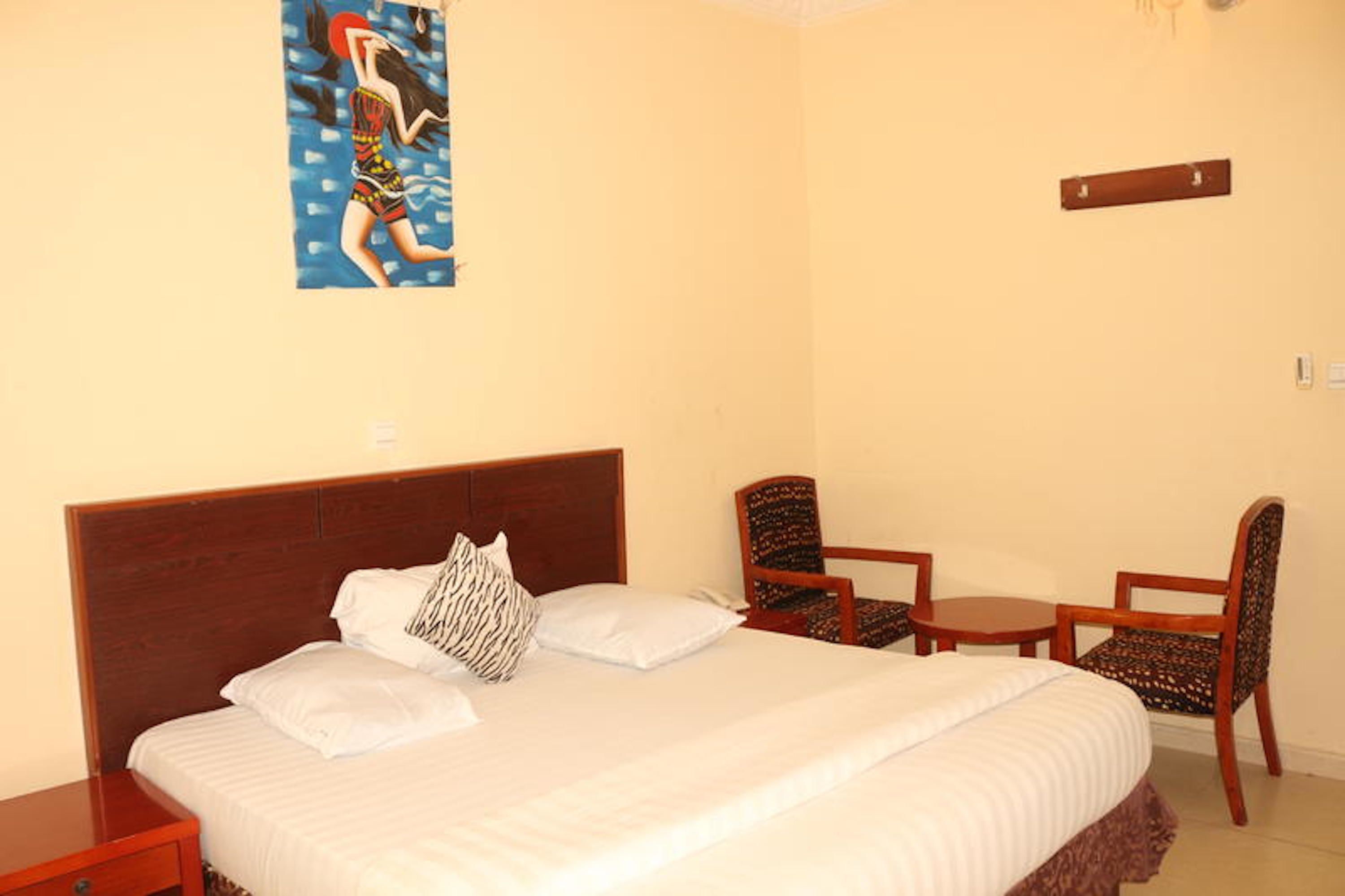 Cama bem arrumada com lençóis brancos e travesseiro estampado, mesa de cabeceira de madeira, duas poltronas estofadas e uma pequena mesa de madeira.