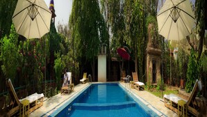 Une piscine entourée de chaises longues en bois et de parasols, avec une végétation luxuriante et un bâtiment traditionnel en arrière-plan.