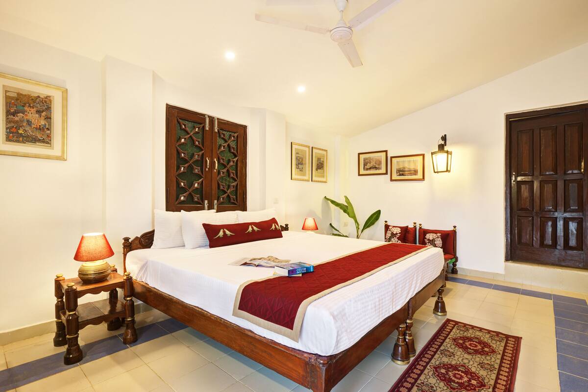 A well-lit bedroom with a large bed, wooden headboard, and a red bedspread. There is a nightstand with a lamp and a wall-mounted shelf.
