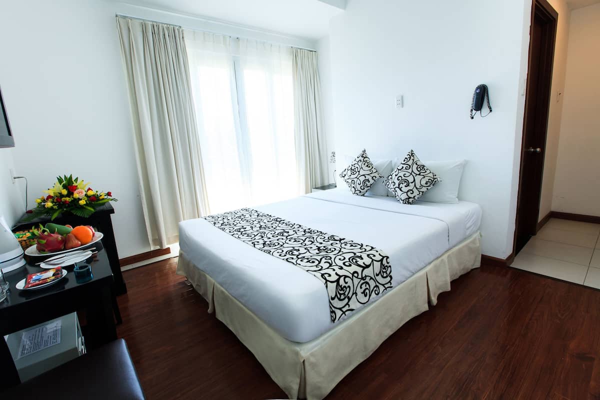 A hotel room with a bed, a desk with a lamp, a fruit bowl, and a window with curtains.