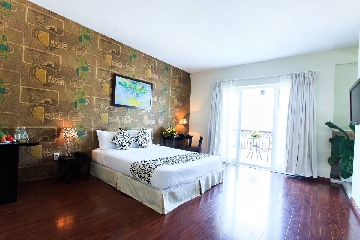 A hotel room with a large bed, a desk with a lamp, a TV, and a balcony with a view.