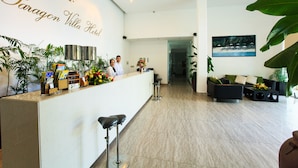 A reception area with a counter, two staff members, a framed picture on the wall, and a seating area with a sofa and chairs. - Paragon Villa Hotel (Nam Nha Trang)