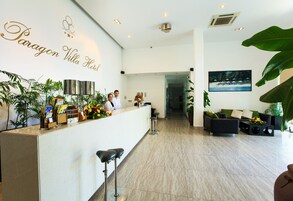 Zona de recepción con mostrador, dos empleados, un cuadro en la pared y un espacio de estar con sofá y sillones.