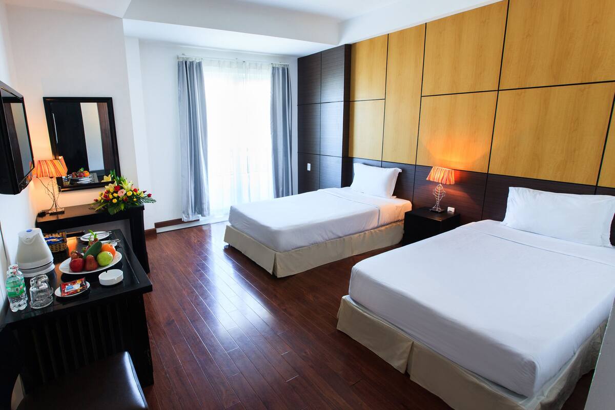 A hotel room with two beds, a desk with a lamp, a mirror, and a window with curtains.