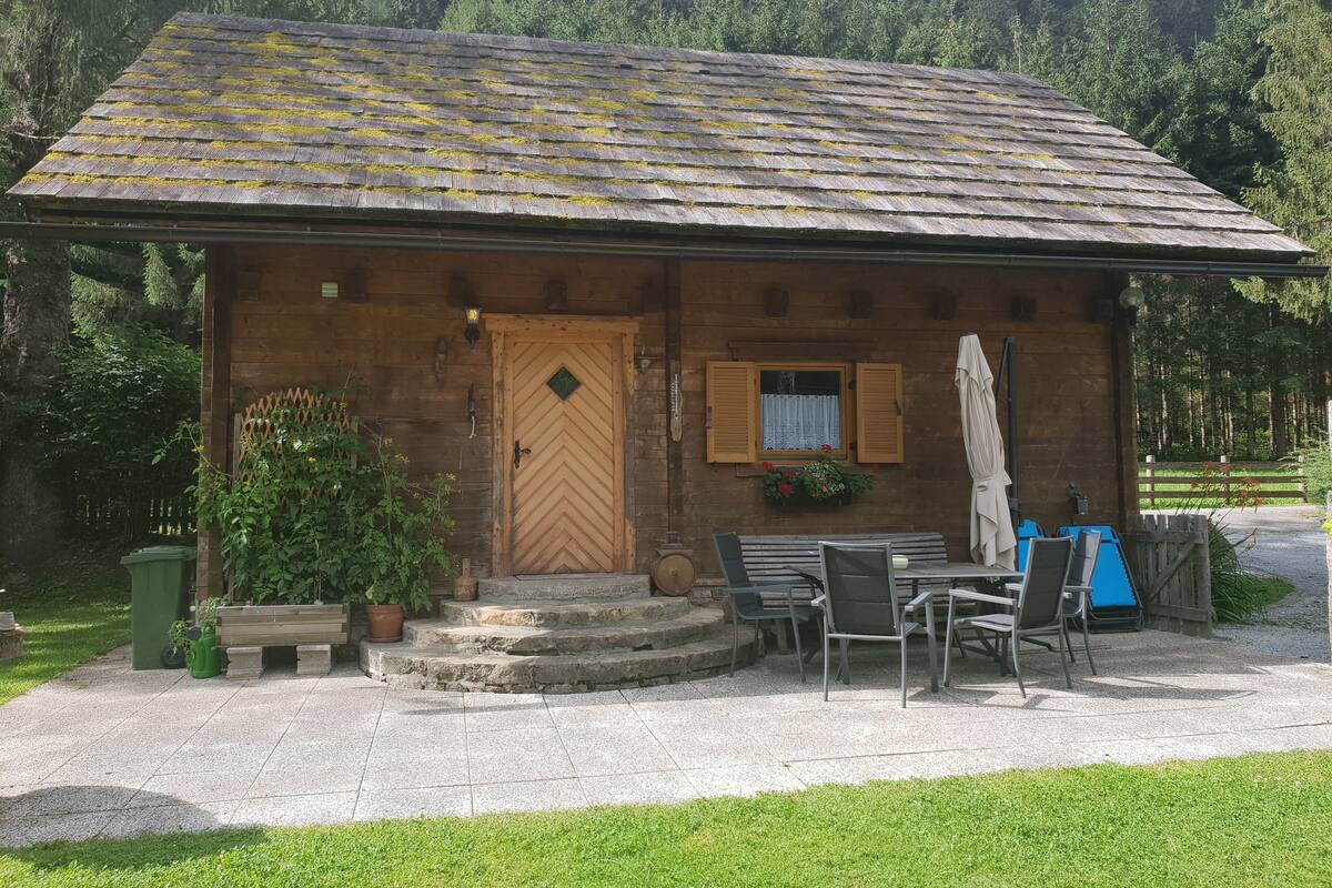 A rustic wooden cabin with a sloped roof, a wooden door, and a small window with shutters. There are outdoor chairs and a table, and a potted plant is visible.