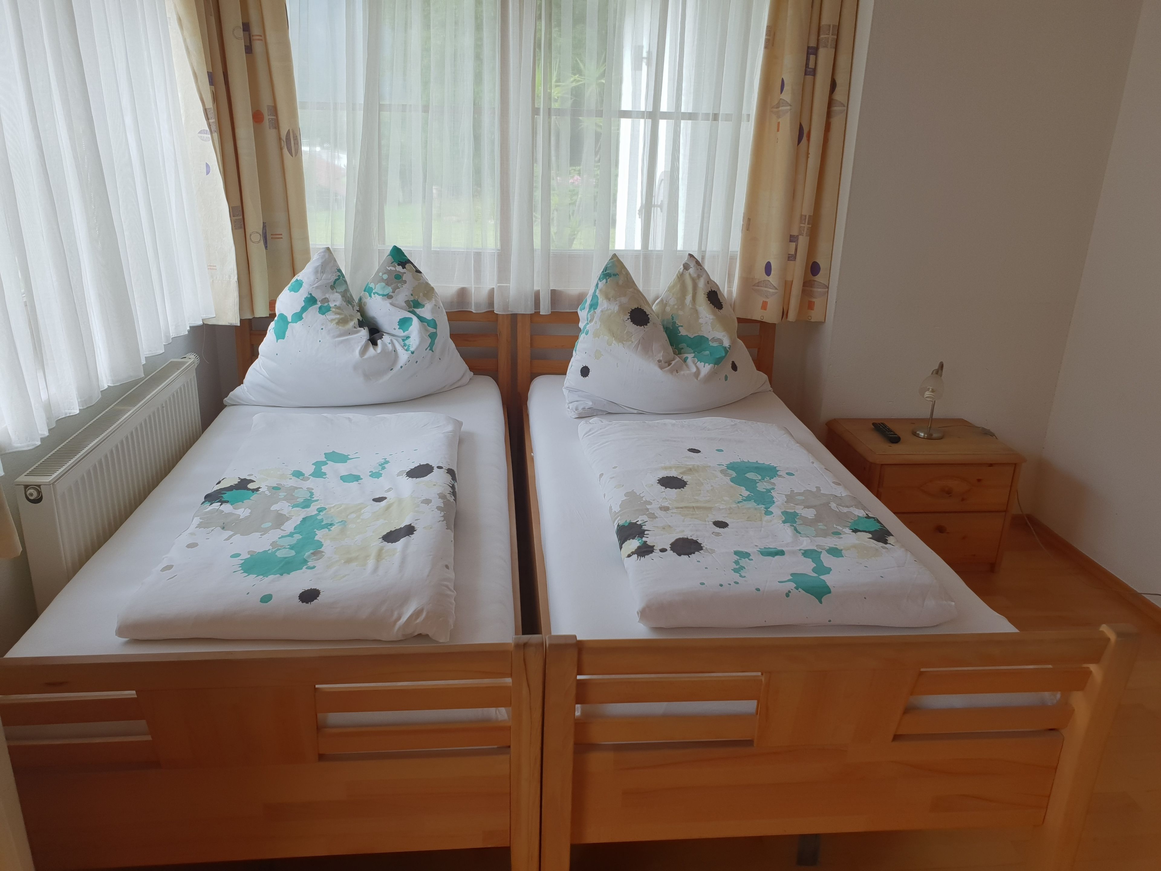Une chambre double avec deux lits en bois, chacun recouvert d’une courtepointe blanche et agrémenté d’oreillers assortis. Une table de chevet en bois, sur laquelle se trouvent une lampe et une télécommande, est placée à côté de l’un des lits.