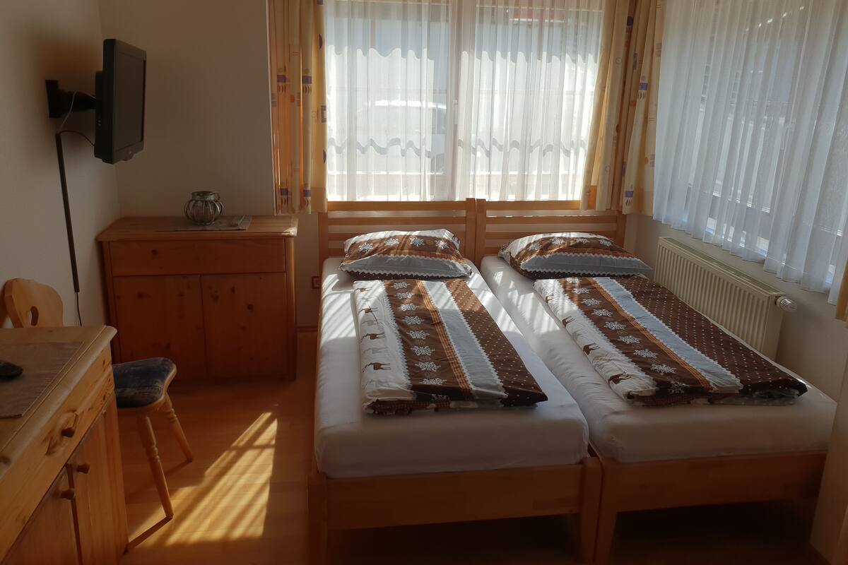 A small, well-lit room with two single beds, a wooden desk, a chair, and a TV mounted on the wall.