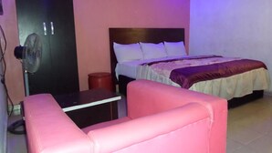 A bedroom with a bed, a bedside table, a chair, a fan, and a red trash bin. - Time View Hotel & Suites (Lagos)