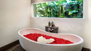 Una bañera llena de flores rojas y blancas, una ventana con plantas y un lavamanos de piedra.