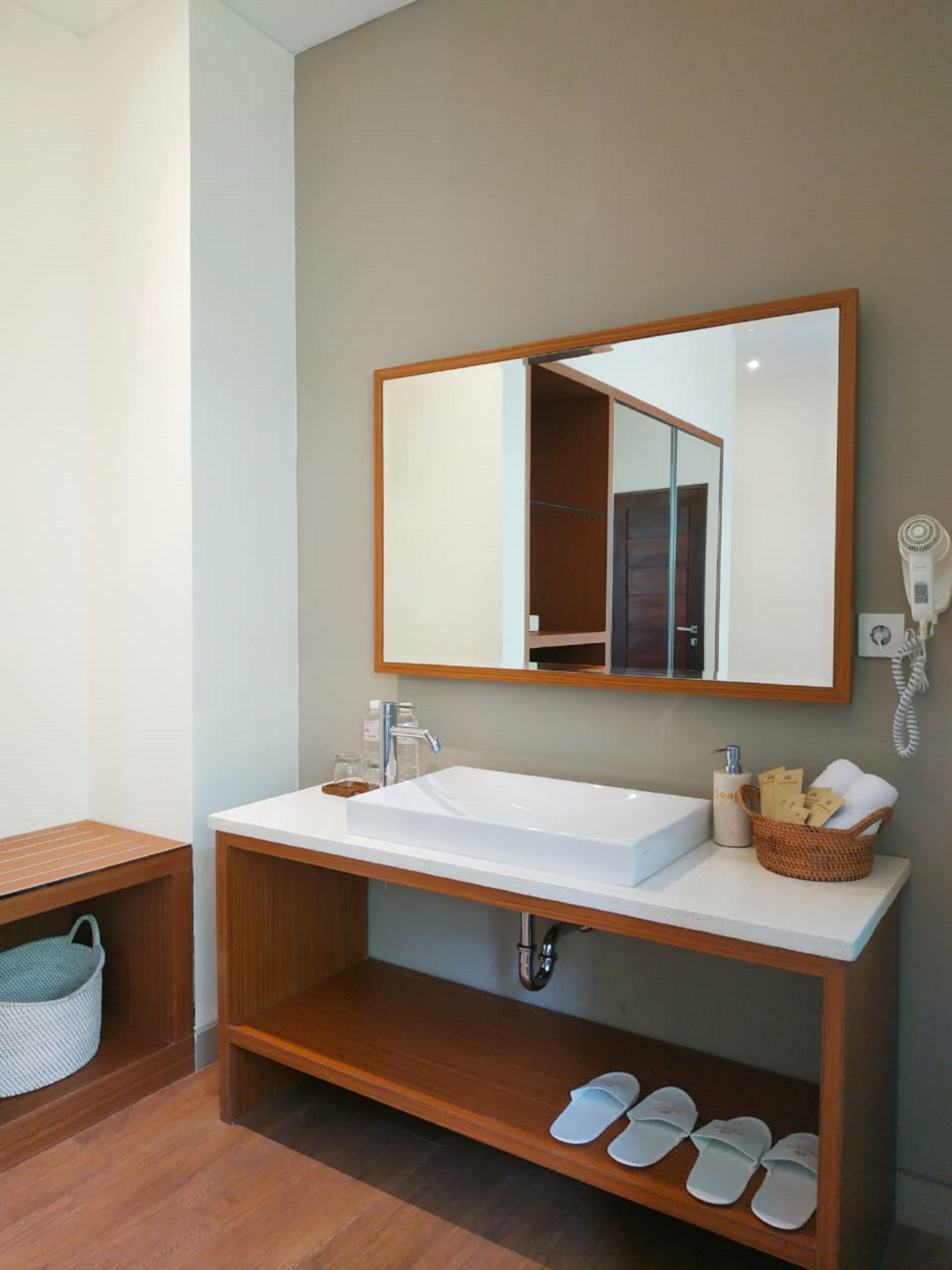 Een badkamer met een witte wastafel, houten wastafelmeubel, een spiegel, een haardroger, een telefoon en een plank met pantoffels.