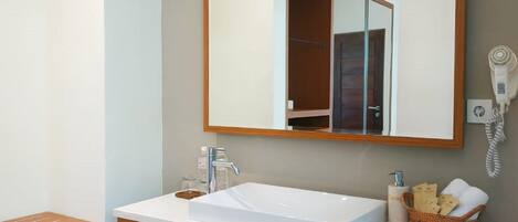 A bathroom with a white sink, wooden vanity, a mirror, a hairdryer, a phone, and a shelf with slippers.
