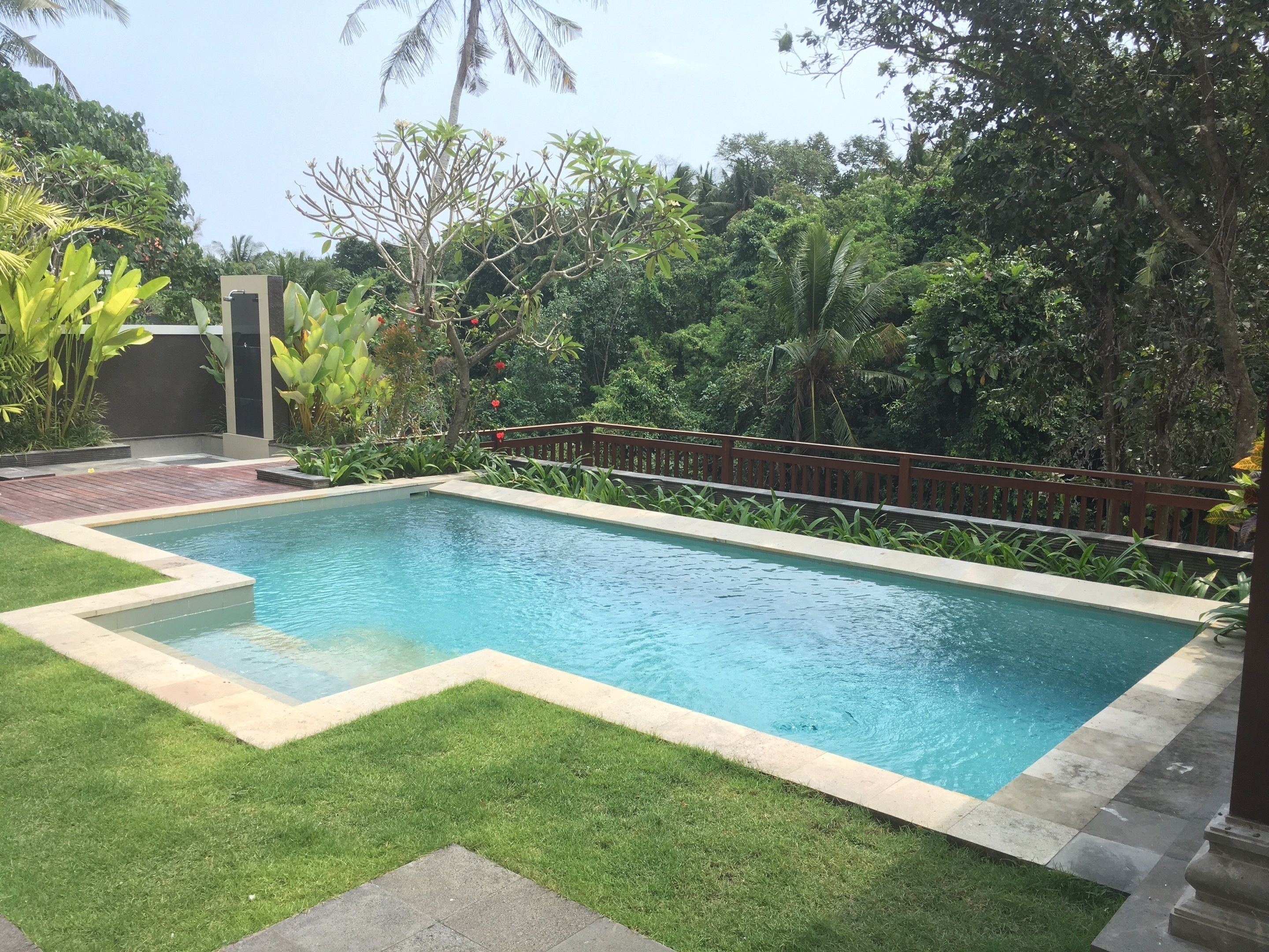 A rectangular swimming pool with a wooden deck and a stone border, surrounded by lush greenery and a wooden fence.