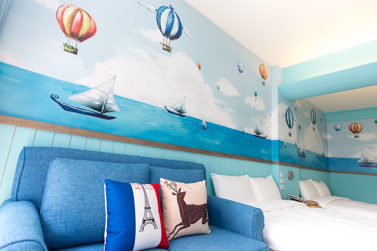 Una habitación con un sofá azul, una cama con ropa de cama blanca y un mural en la pared que muestra globos aerostáticos y veleros.