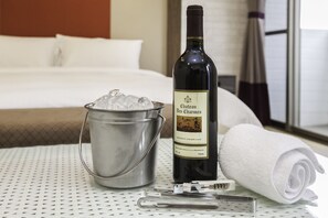 Une chambre d’hôtel avec un lit, une bouteille de vin Château des Charmes, un seau en métal rempli de glace et une serviette blanche roulée posée sur une table.