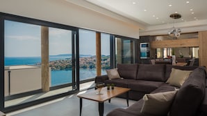 A modern living room with a large window, a sofa, a coffee table, and a kitchen area visible through the glass. - Trogir Pearl Luxury Resort (Okrug)