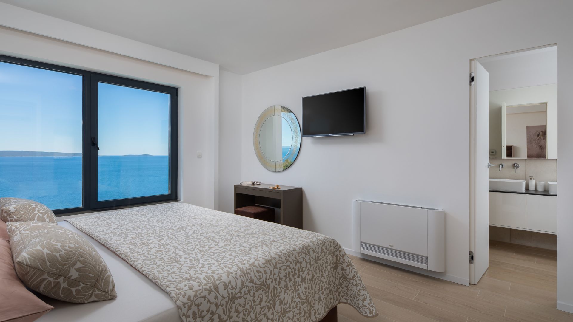 Een slaapkamer met een groot raam, een bed met beddengoed met patroon, een nachtkastje, een ronde spiegel, een televisie, en uitzicht op zee.