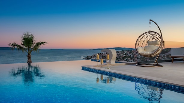 A infinity pool with clear blue water, a palm tree, a hanging chair, and a view of the sea and distant hills. - Trogir Pearl Luxury Resort (Okrug)