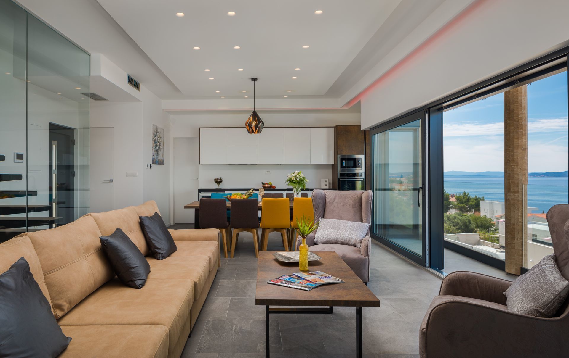 A modern living room with a sofa, coffee table, and chairs, a dining area with a table and chairs, and a kitchen with cabinets and appliances.