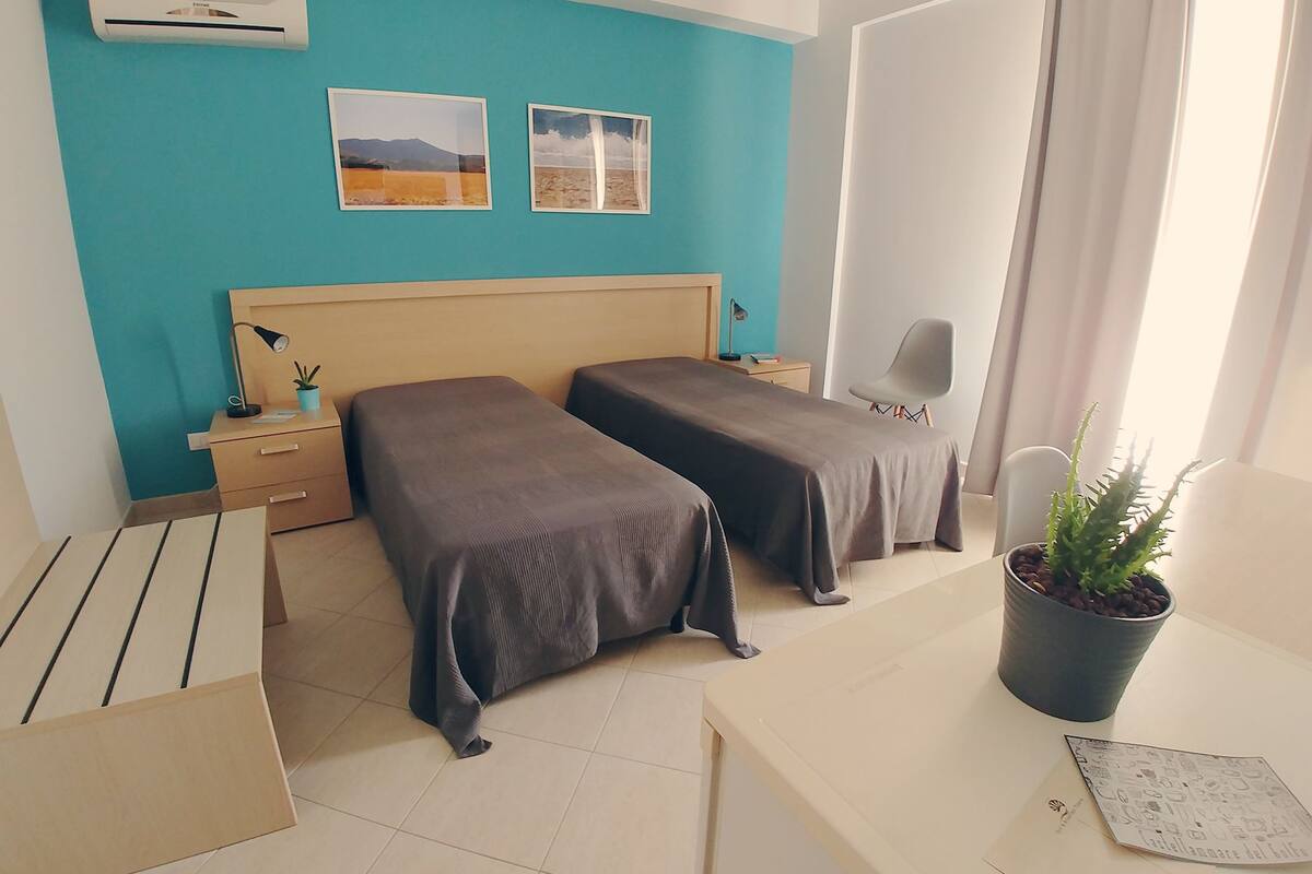 A hotel room with two single beds, a desk with a chair, a potted plant, and two framed pictures on the wall.