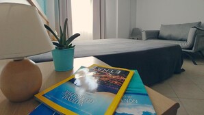 A hotel room with a bed, a desk with a lamp and books, a potted plant, and a view of the city through the window.