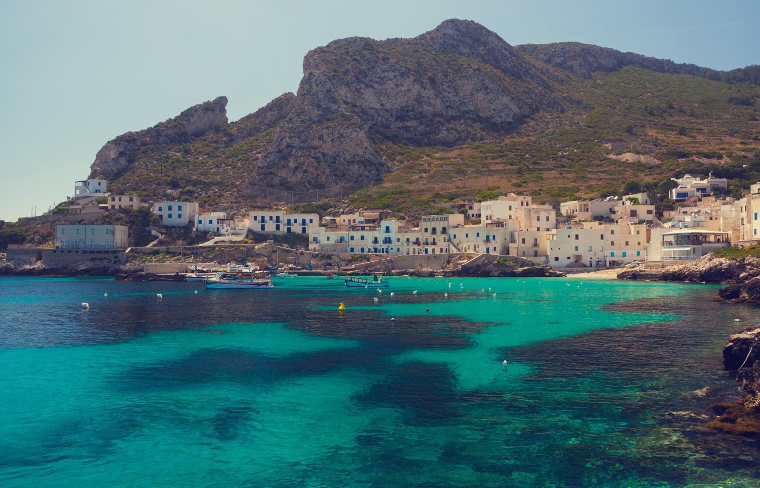 Une ville côtière avec des bâtiments qui bordent le front de mer, une eau turquoise et limpide, et une montagne rocheuse en arrière-plan.