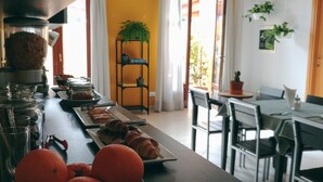 A dining area with a long table set with pastries and oranges, a black metal shelf with plants, and a view of the outdoors through a door.