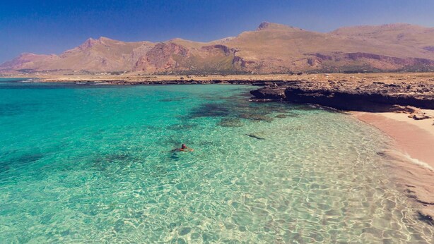 A beach with clear turquoise water, a person swimming, and mountains in the background.