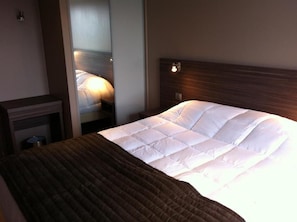 A hotel room with a bed, a nightstand, a mirror, and a wall-mounted light fixture.