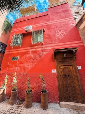 A red building with a wooden door and windows, potted plants in front, and a blue sky in the background.