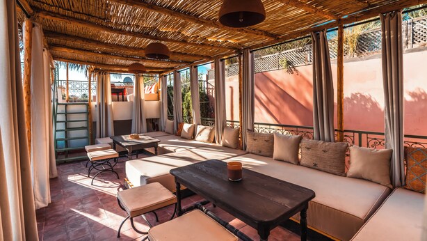 A covered outdoor seating area with cushioned benches and tables, large windows, and a thatched roof. - Riad Dar Tiflet (Marrakech)