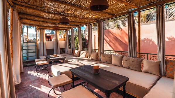 A covered outdoor seating area with cushioned benches and tables, large windows, and a thatched roof. - Riad Dar Tiflet (Marrakech)
