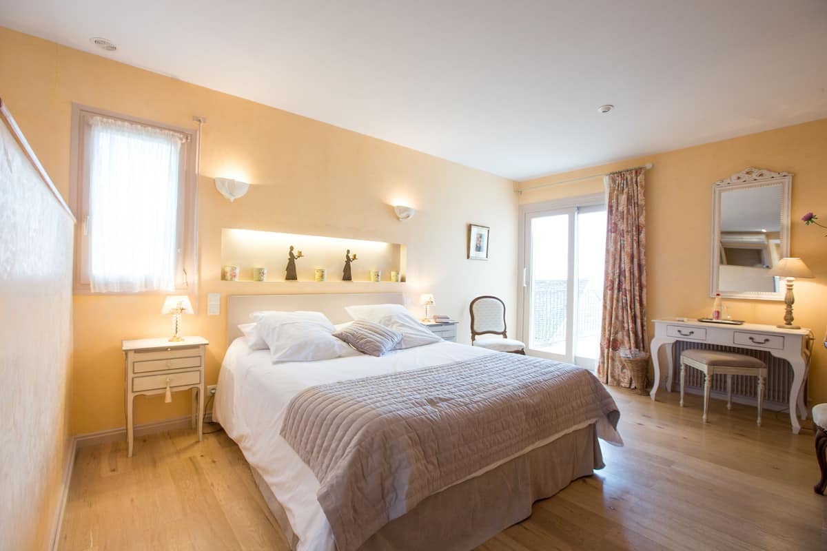 Ein Schlafzimmer mit Bett, Nachttischen, einem Schminktisch, einem Sessel und einem Fenster mit Vorhängen.