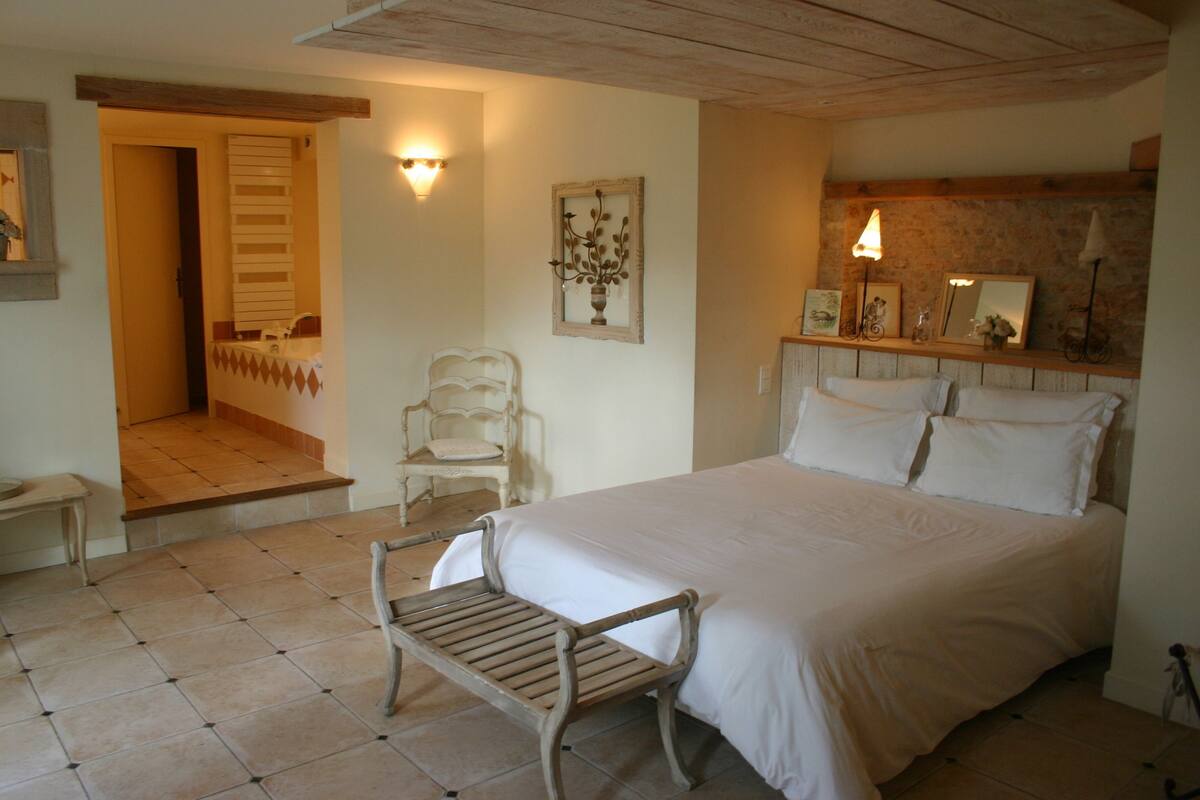 Ein Schlafzimmer mit einem großen Bett, einer hölzernen Bank, einem Sessel und einem kleinen Tisch. Eine Tür führt in einen anderen Raum, und an der Wand befindet sich ein Regal mit Dekorationen.