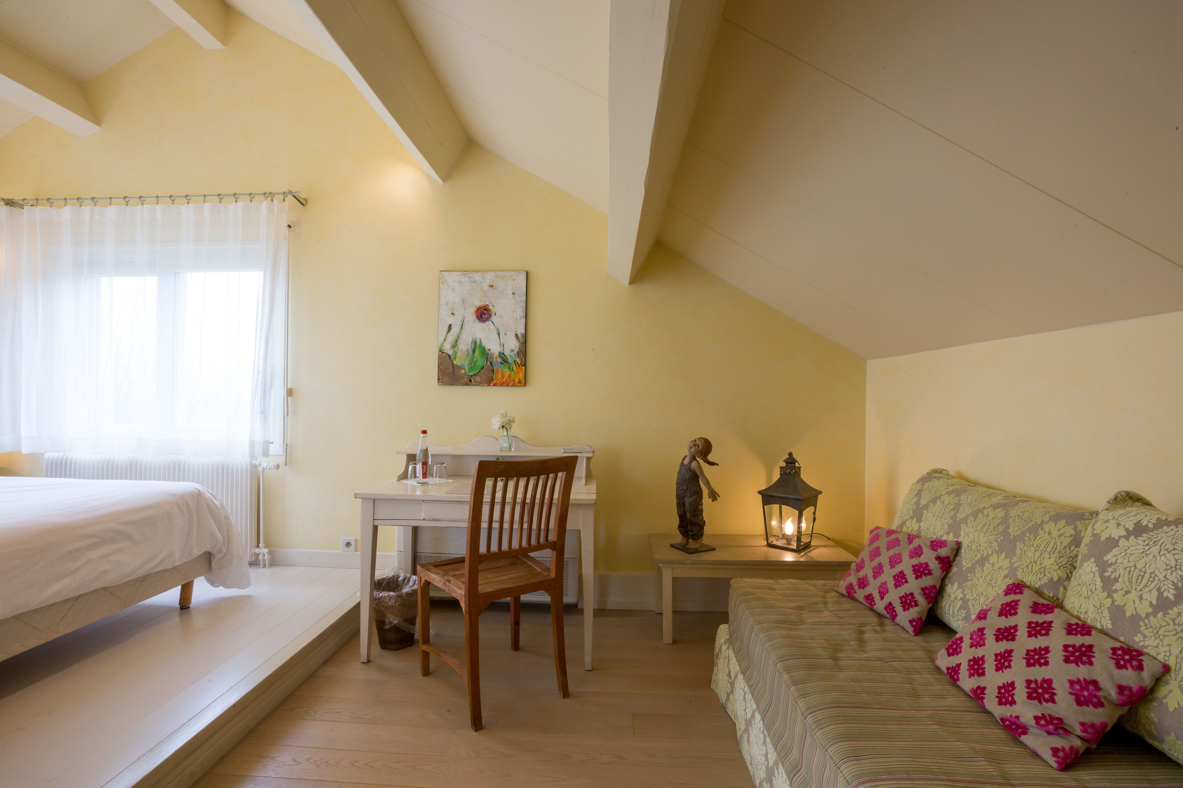 A bedroom with a bed, a desk, a chair, a small table with a statue, and a sofa with patterned cushions.
