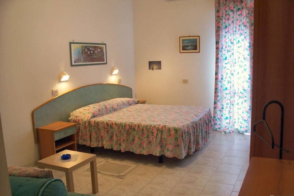 Une petite pièce agréable et confortable, avec un lit, un bureau et une chaise. On y trouve deux lampes fixées au mur et une fenêtre ornée de rideaux à motifs floraux.
