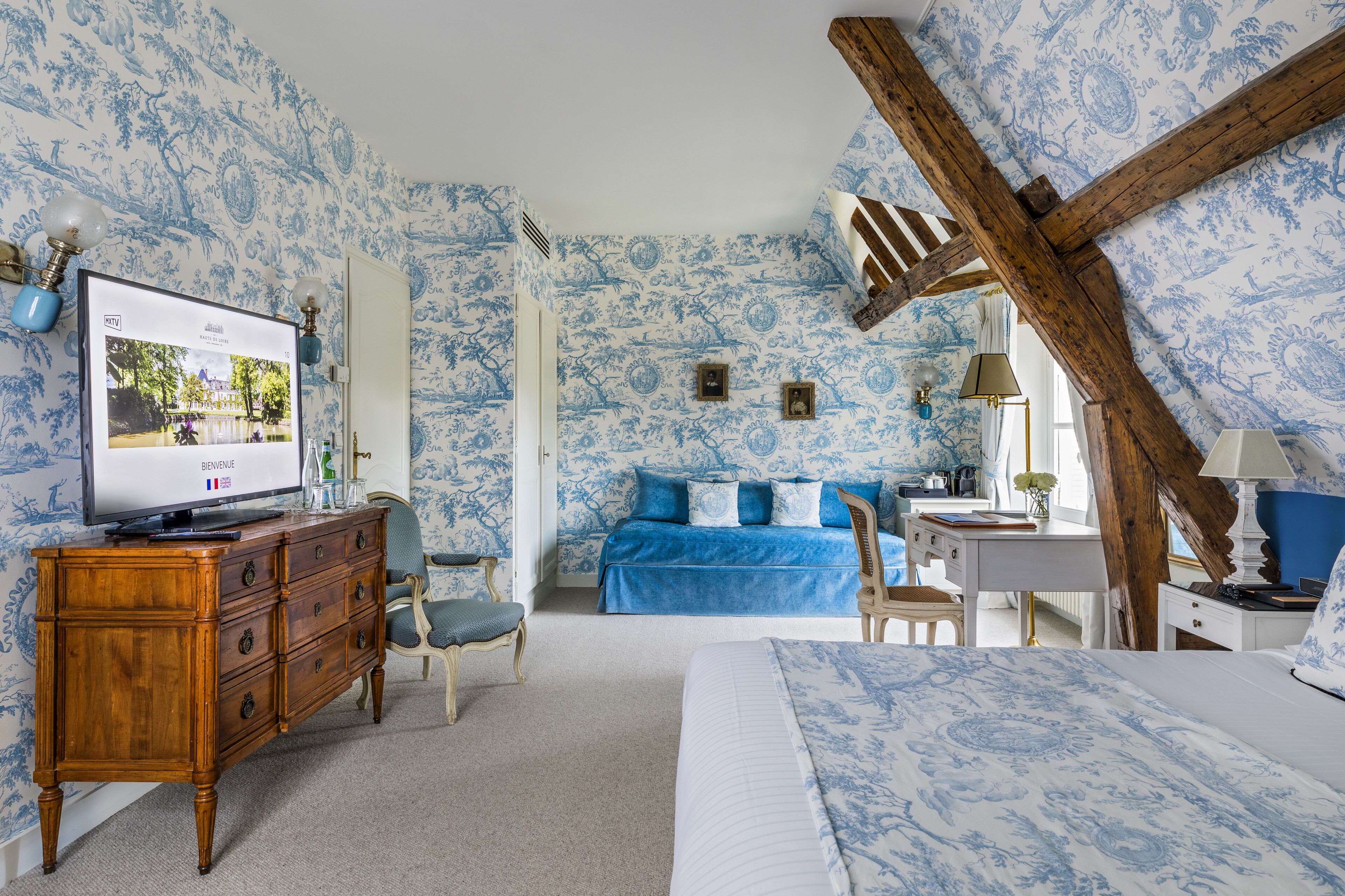 A bedroom with a TV, wooden dresser, blue bed, and a wooden ceiling beam.