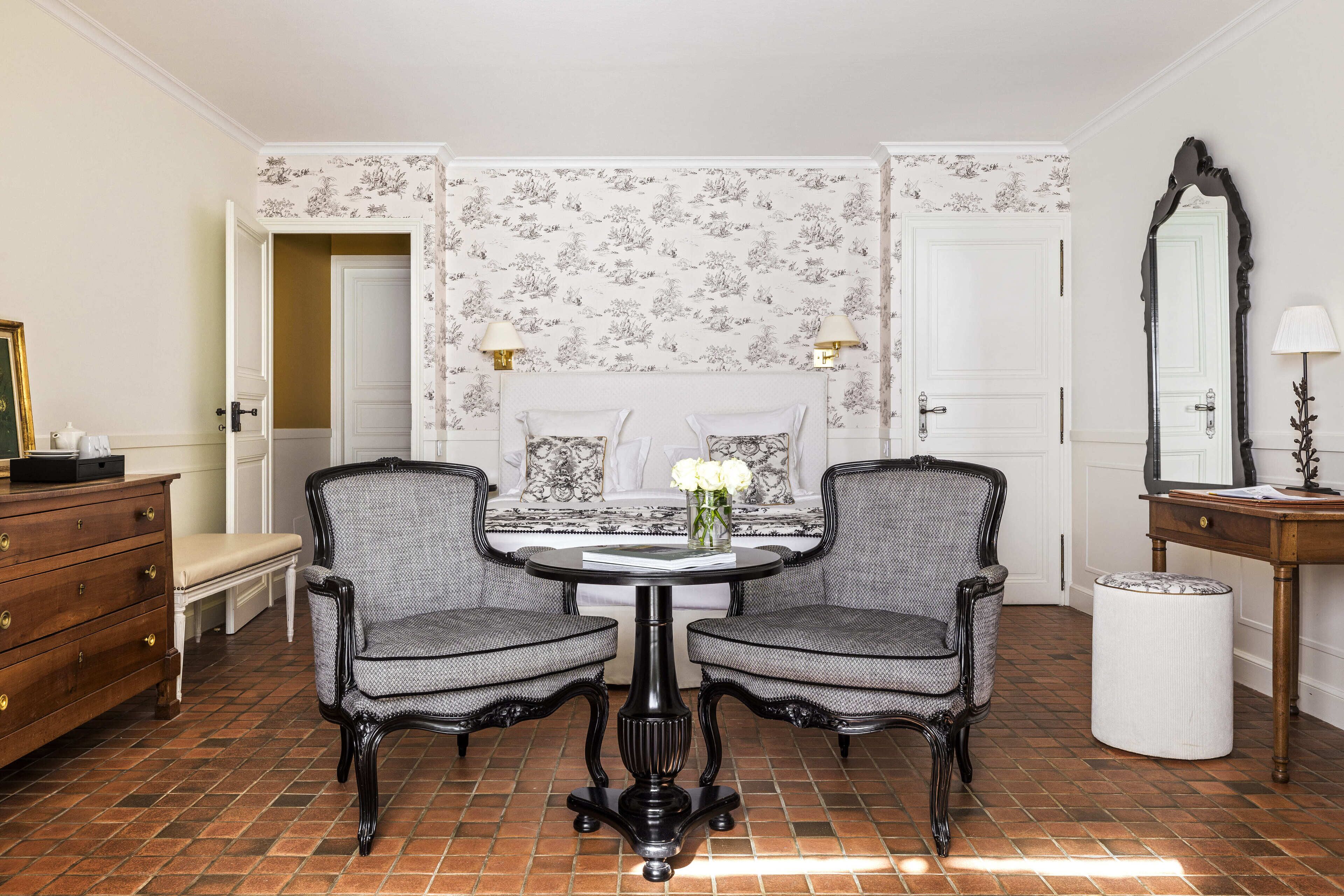 A room with a round table, two chairs, a bench, a dresser, a mirror, and patterned wallpaper.