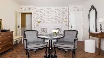 A room with a round table, two chairs, a bench, a dresser, a mirror, and patterned wallpaper.
