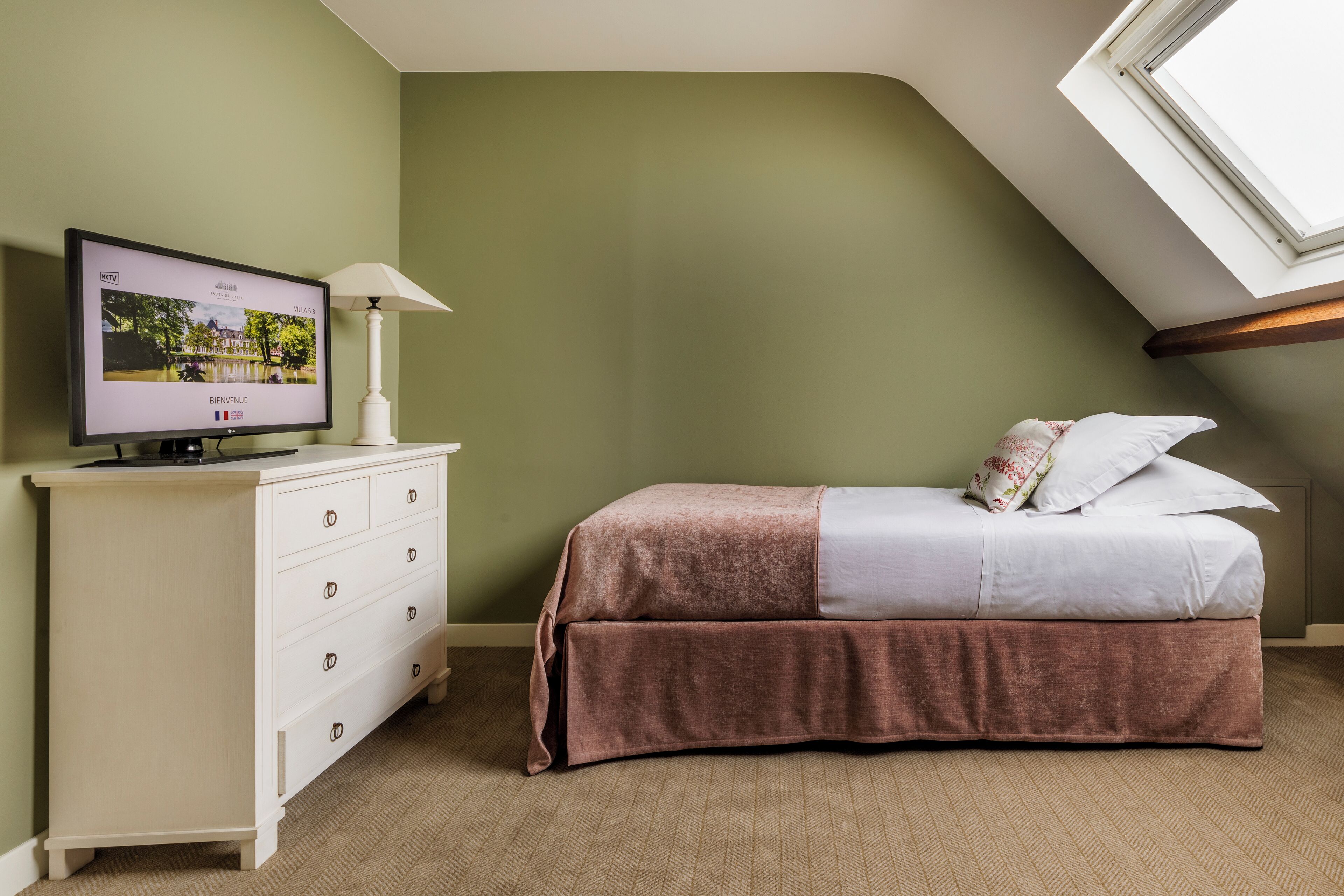 Un dormitorio con televisor sobre un aparador, una cama con cubrecama marrón y un tragaluz.
