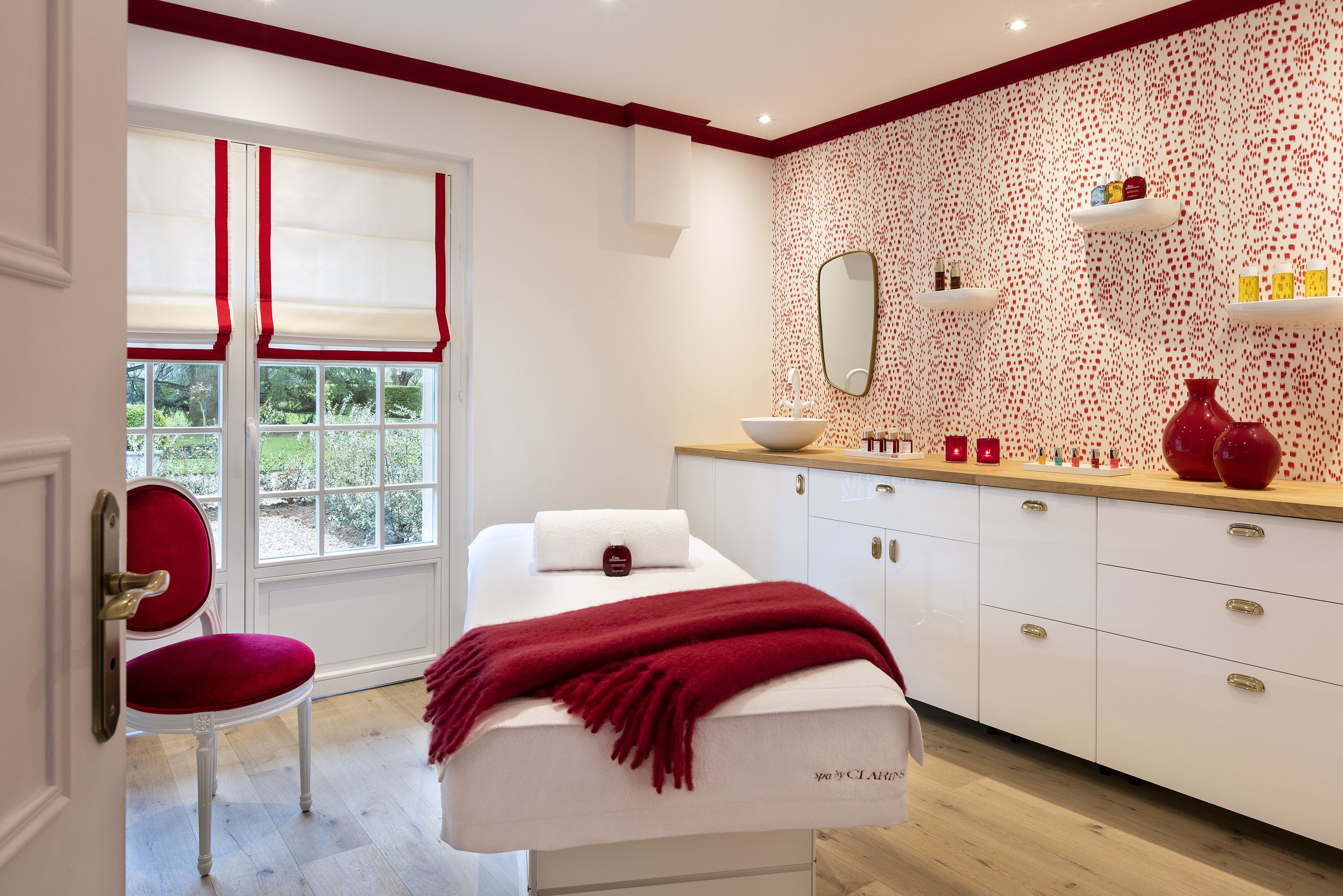Une salle de spa équipée d’une table de massage, d’un fauteuil rouge, d’une fenêtre avec des stores rouges, d’un miroir et d’un meuble en bois contenant des articles de toilette.