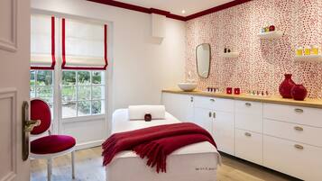 Une salle de spa équipée d’une table de massage, d’un fauteuil rouge, d’une fenêtre avec des stores rouges, d’un miroir et d’un meuble en bois contenant des articles de toilette.
