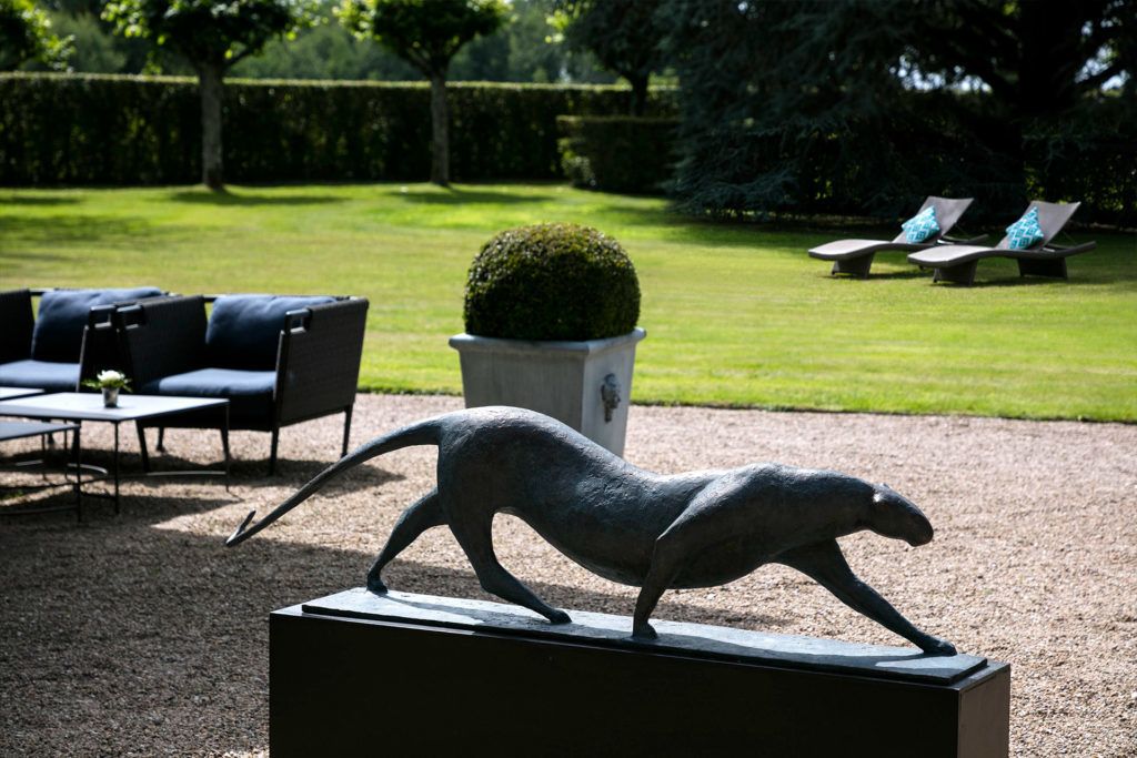 A sculpture of a wild cat on a pedestal in a garden with a lawn, seating area, and hedges.